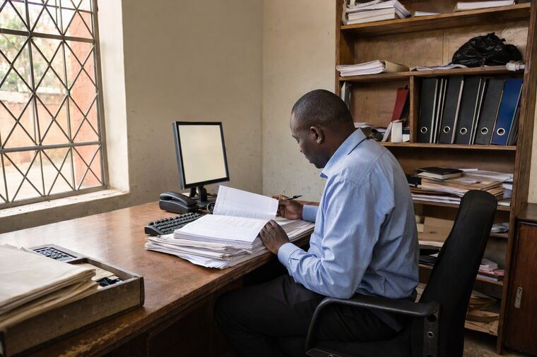 A manager working from a paper ledger beside an unused monitor